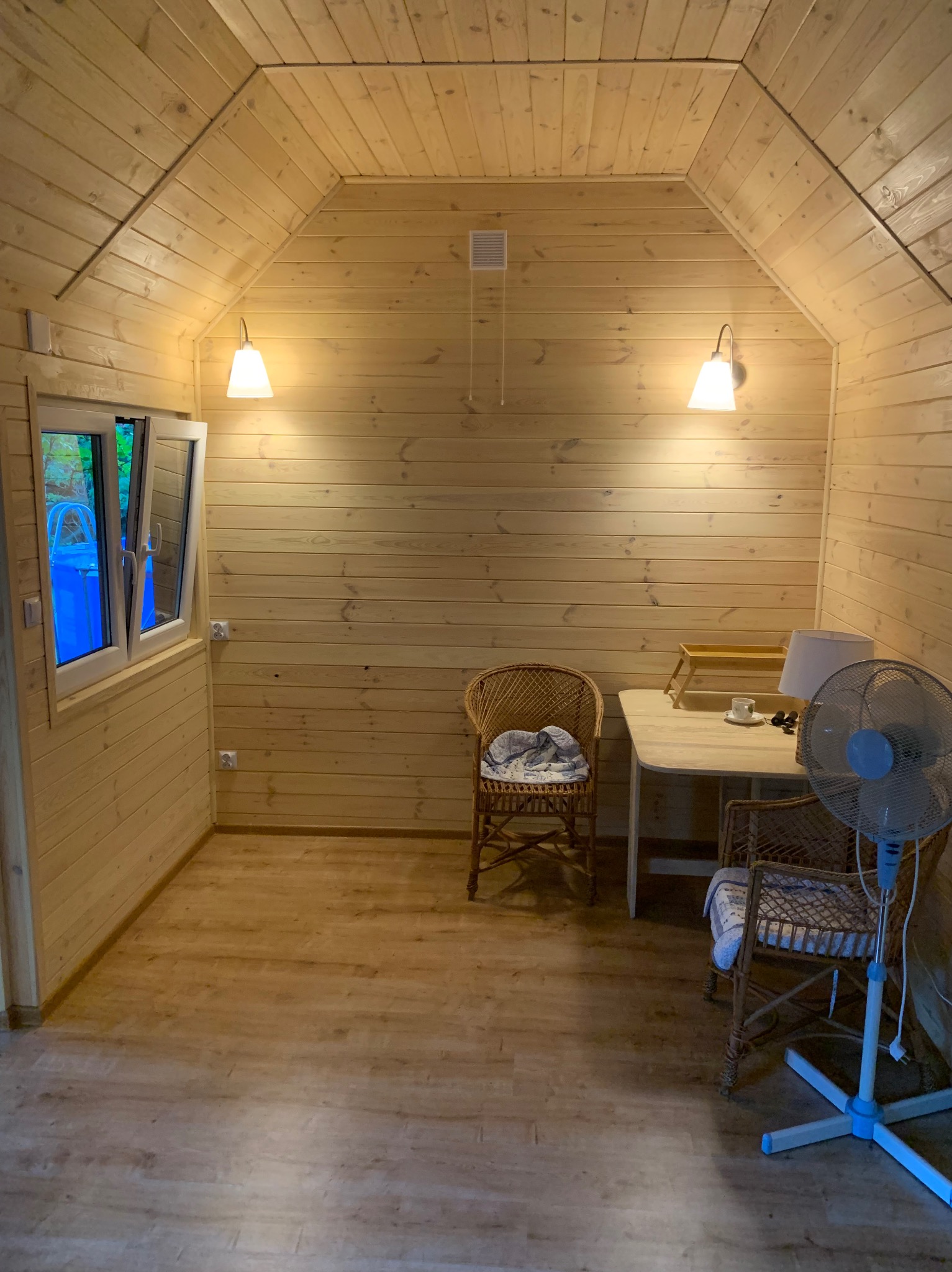 Wnętrze domku z drewnianą boazerią, oświetlone lampami ściennymi. W środku wiklinowe krzesła, stół z lampką i wentylator. Widoczne okno z otwartym skrzydłem.