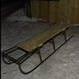 Paweł Kulaszewicz - Metalowe sanki z drewnianym siedziskiem stoją na śniegu przed drewnianymi drzwiami z metalowymi zawiasami w słabym oświetleniu.