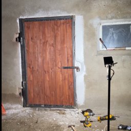 Paweł Kulaszewicz - Drewniane drzwi z metalową ramą w surowym otoczeniu, widoczne narzędzia i lampa warsztatowa. Wykończone drzwi w trakcie montażu, oświetlone lampą.