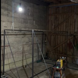 Paweł Kulaszewicz - Konstrukcja metalowa w trakcie spawania w warsztacie. Widoczny spawacz i elementy konstrukcyjne. Surowe wnętrze z ceglanymi ścianami i drewnianym stropem.