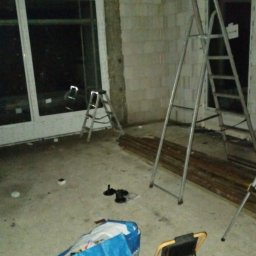 KUKIS INSTALL SZYMON KUKIEŁKA - Wnętrze w trakcie remontu: okno PCV, dwie drabiny aluminiowe, torba narzędziowa i lampa budowlana na podłodze. Surowy stan ścian z betonu komórkowego.