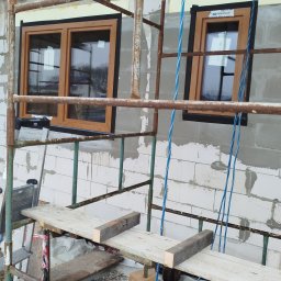 KUKIS INSTALL SZYMON KUKIEŁKA - Montaż okien z drewnianą ramą w surowym murze z białych bloczków, widoczne rusztowanie i niebieskie liny. Etap prac wykończeniowych na budowie.