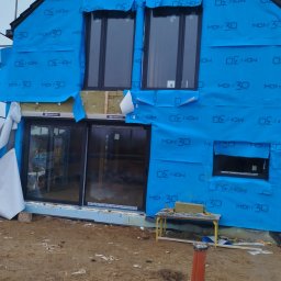 KUKIS INSTALL SZYMON KUKIEŁKA - Dom w trakcie budowy, zabezpieczony niebieską folią izolacyjną. Widoczne okna, fragment ocieplenia i narzędzia. Prace wykończeniowe w toku.