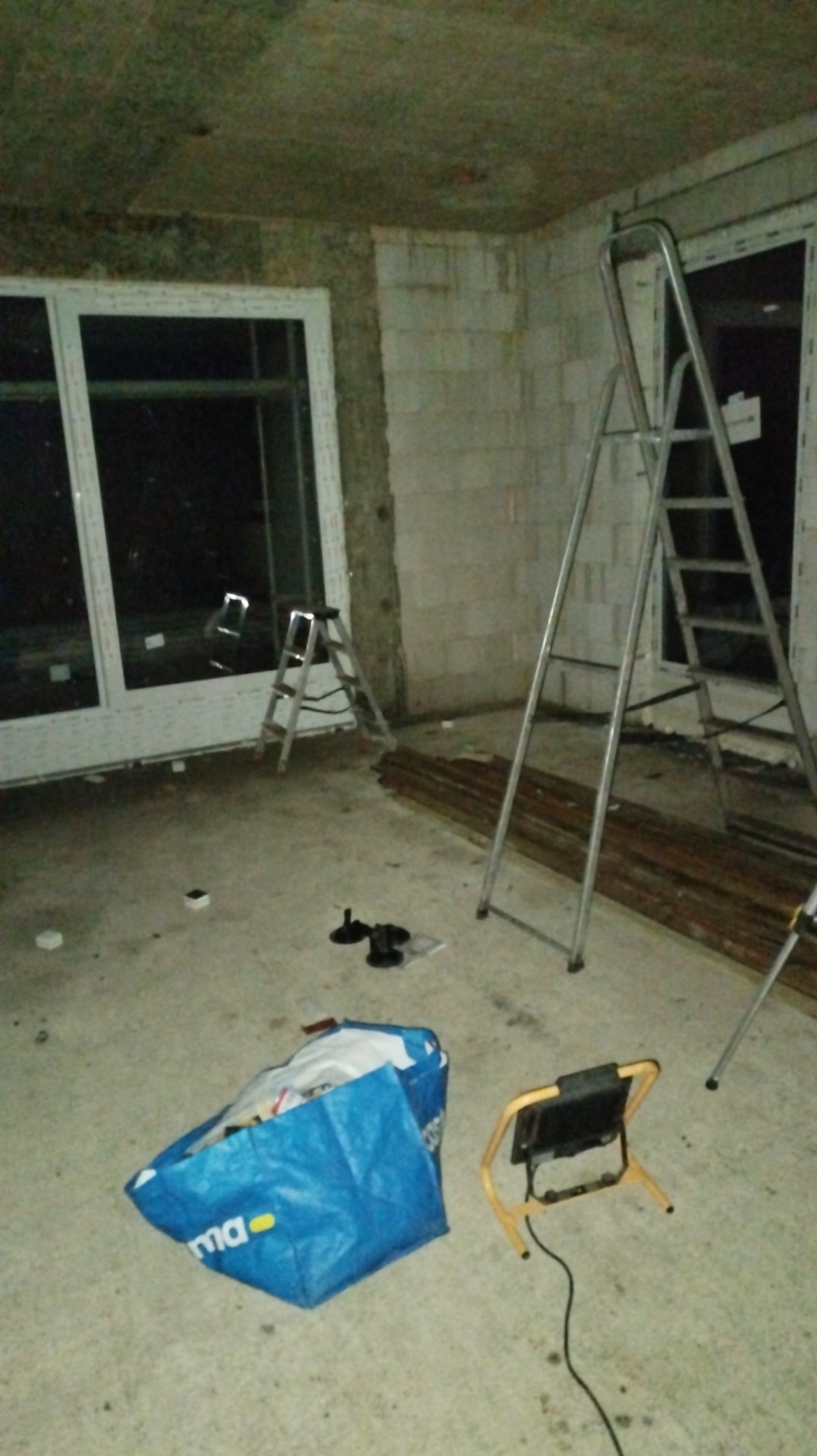 Wnętrze w trakcie remontu: okno PCV, dwie drabiny aluminiowe, torba narzędziowa i lampa budowlana na podłodze. Surowy stan ścian z betonu komórkowego.