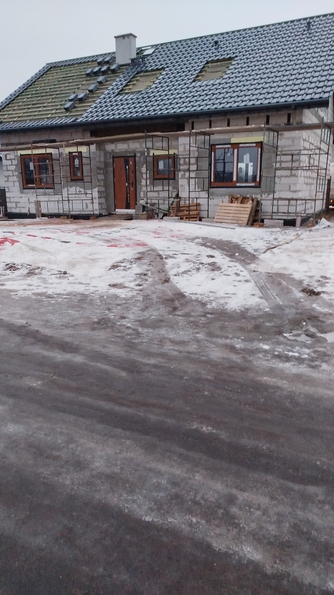 Surowy stan domu w budowie z częściowo położonym dachem z dachówki, widoczne okna i drzwi, teren wokół pokryty śniegiem. Ujęcie z dołu.