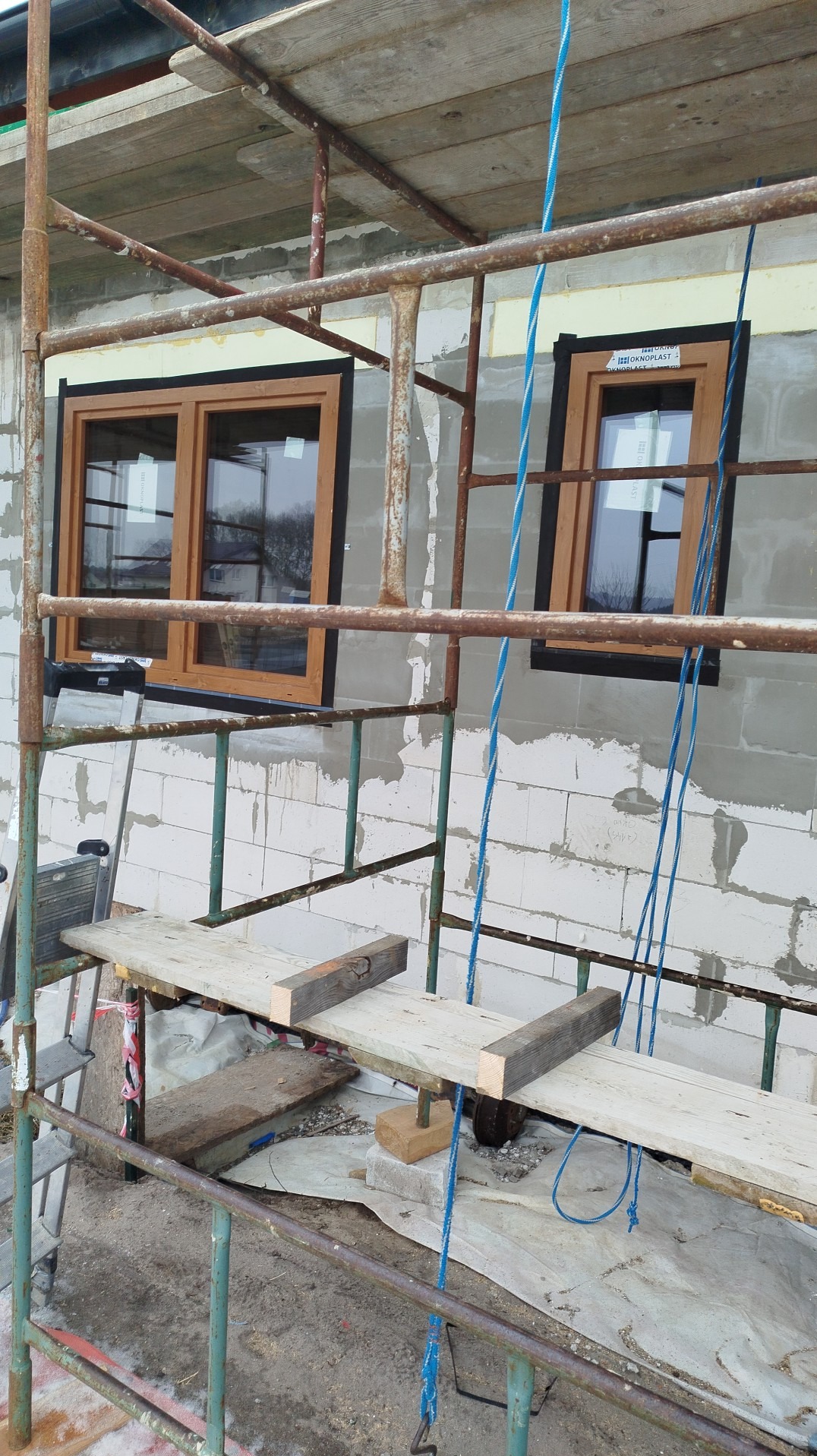 Montaż okien z drewnianą ramą w surowym murze z białych bloczków, widoczne rusztowanie i niebieskie liny. Etap prac wykończeniowych na budowie.