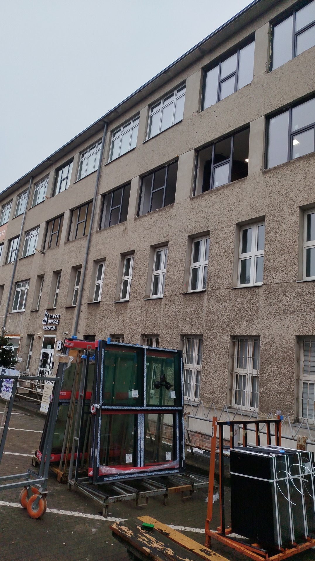 Budynek z częściowo wymienionymi oknami. Na pierwszym planie szyby okienne na wózkach transportowych. Widoczne ślady prac montażowych.