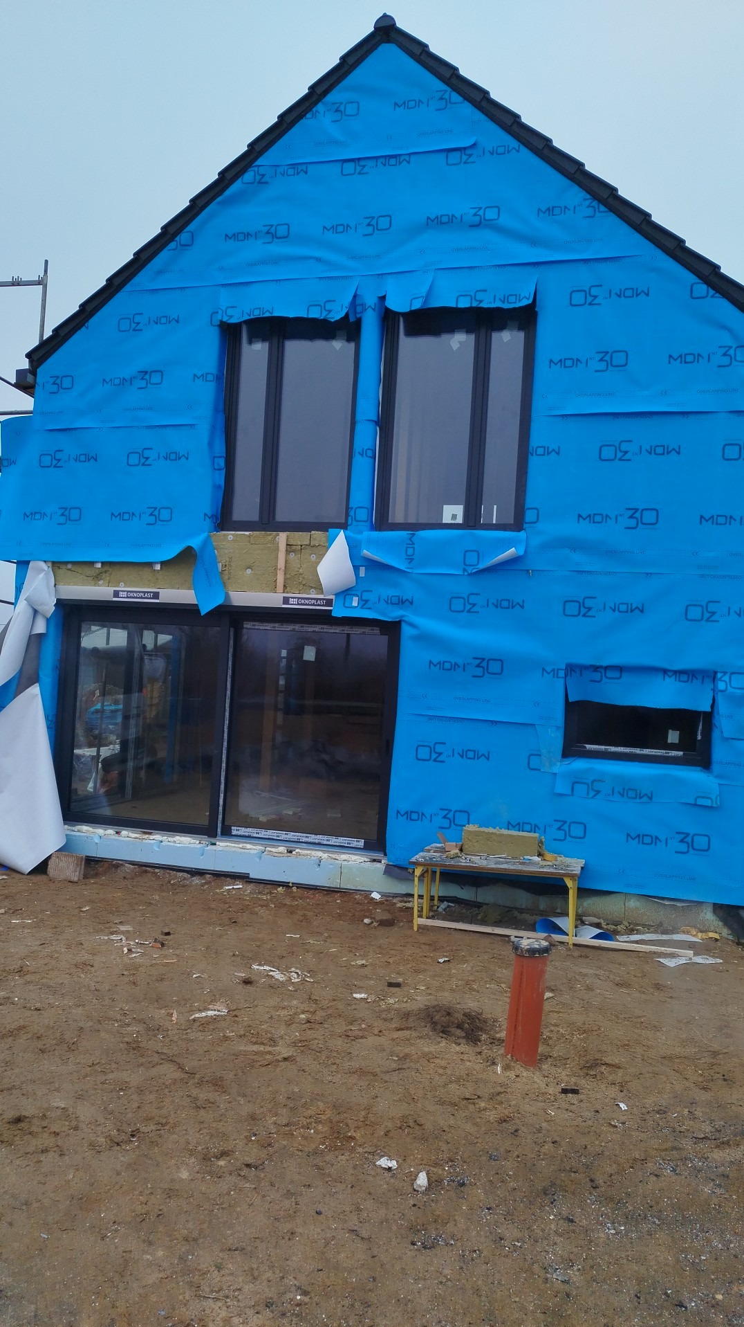 Dom w trakcie budowy, zabezpieczony niebieską folią izolacyjną. Widoczne okna, fragment ocieplenia i narzędzia. Prace wykończeniowe w toku.