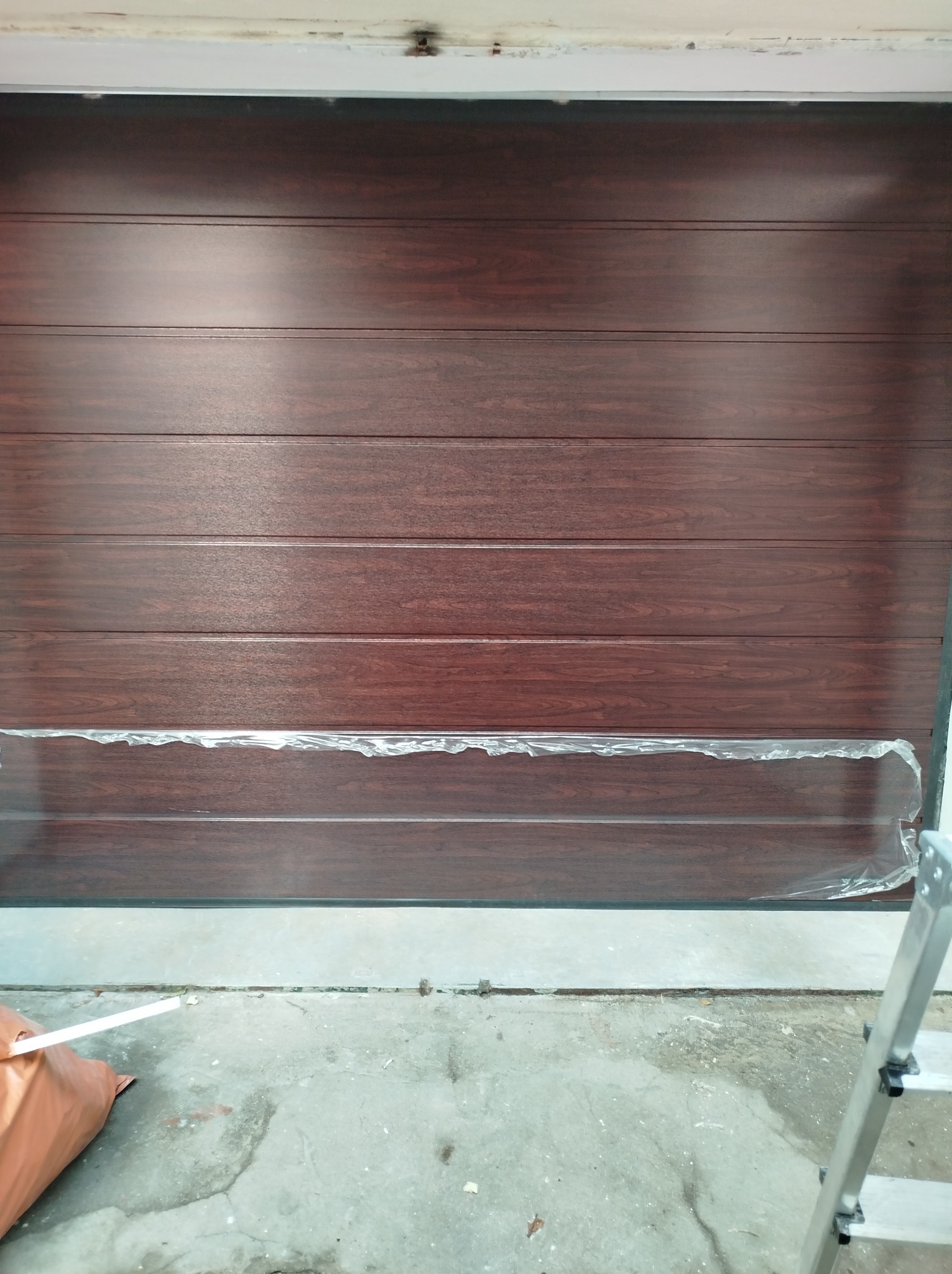 Montaż brązowej, segmentowej bramy garażowej z folią ochronną na dolnym panelu. Widoczna drabina i worek z narzędziami. Ujęcie z bliska, podkreślające detale.