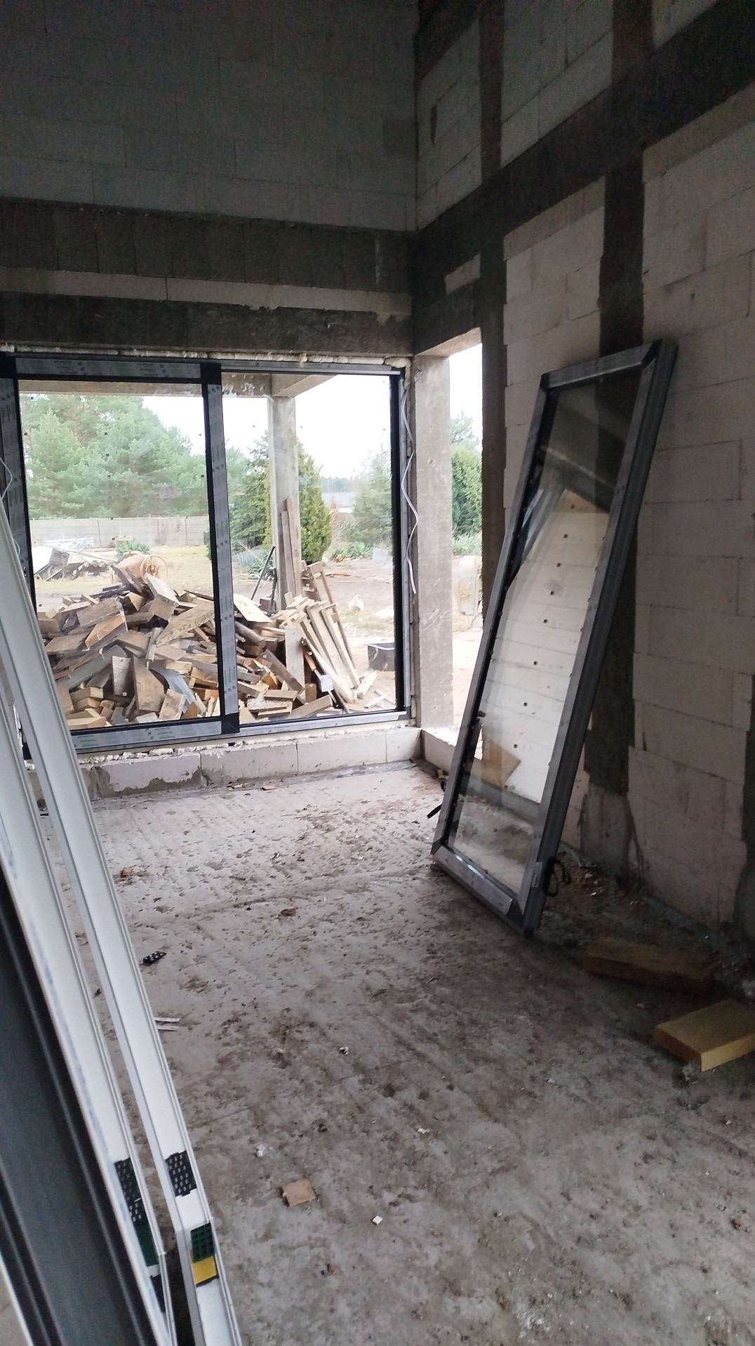 Niezamontowane okno z widokiem na stertę drewna w surowym wnętrzu budynku w trakcie budowy. Okno oparte o ścianę z betonu komórkowego.