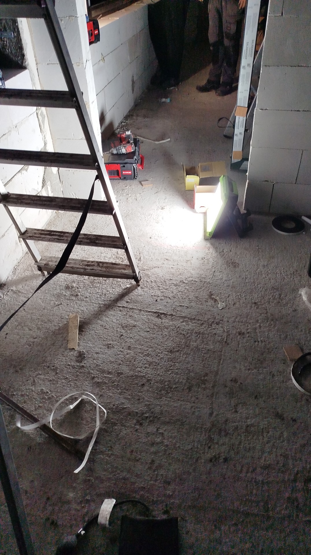 Wnętrze w trakcie prac: drabina, narzędzia akumulatorowe, lampa budowlana oświetlają surowy beton i fragment ściany z bloczków. Widoczne osoby w tle.