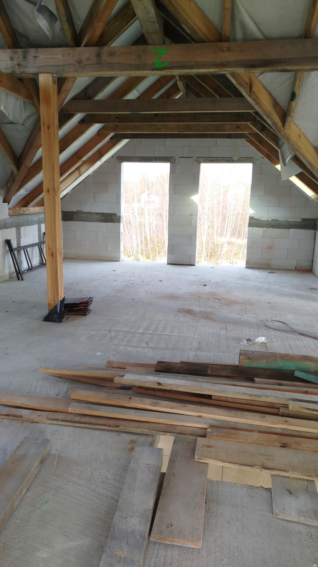 Surowe poddasze z drewnianą konstrukcją dachu, widoczne bloczki betonowe ścian i przygotowane deski podłogowe. Dwa okna z widokiem na drzewa w tle.