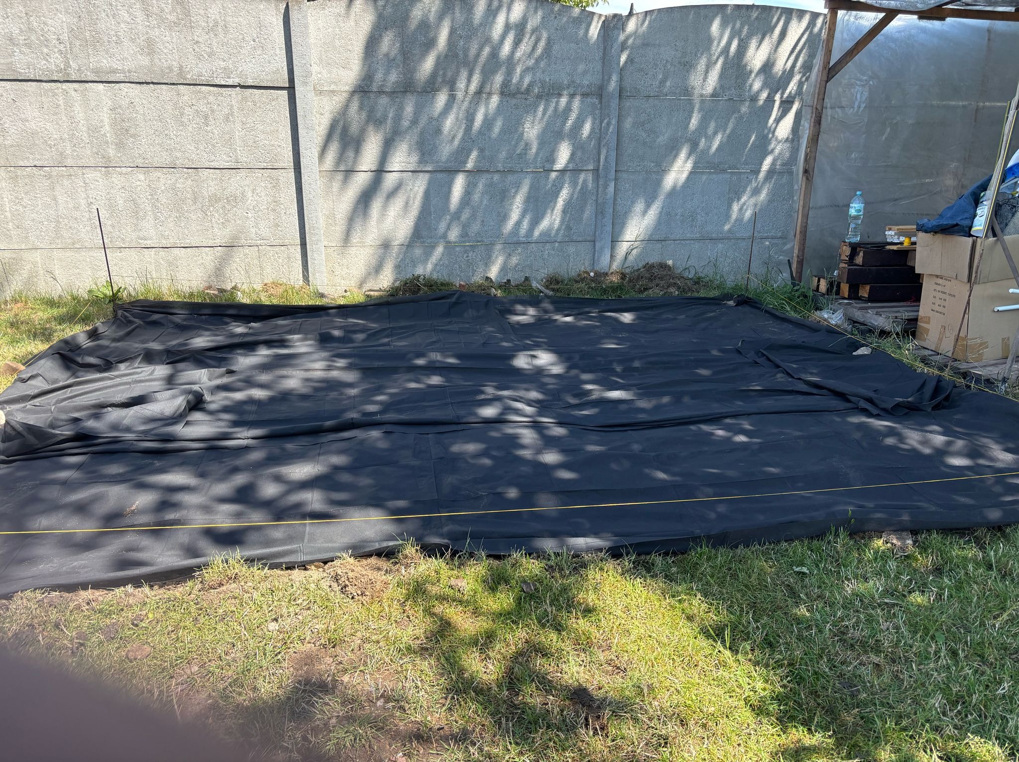 Rozłożona czarna agrowłóknina na trawie, zabezpieczona sznurkiem, przygotowana pod budowę. W tle betonowy płot i prowizoryczna wiata z materiałami budowlanymi.