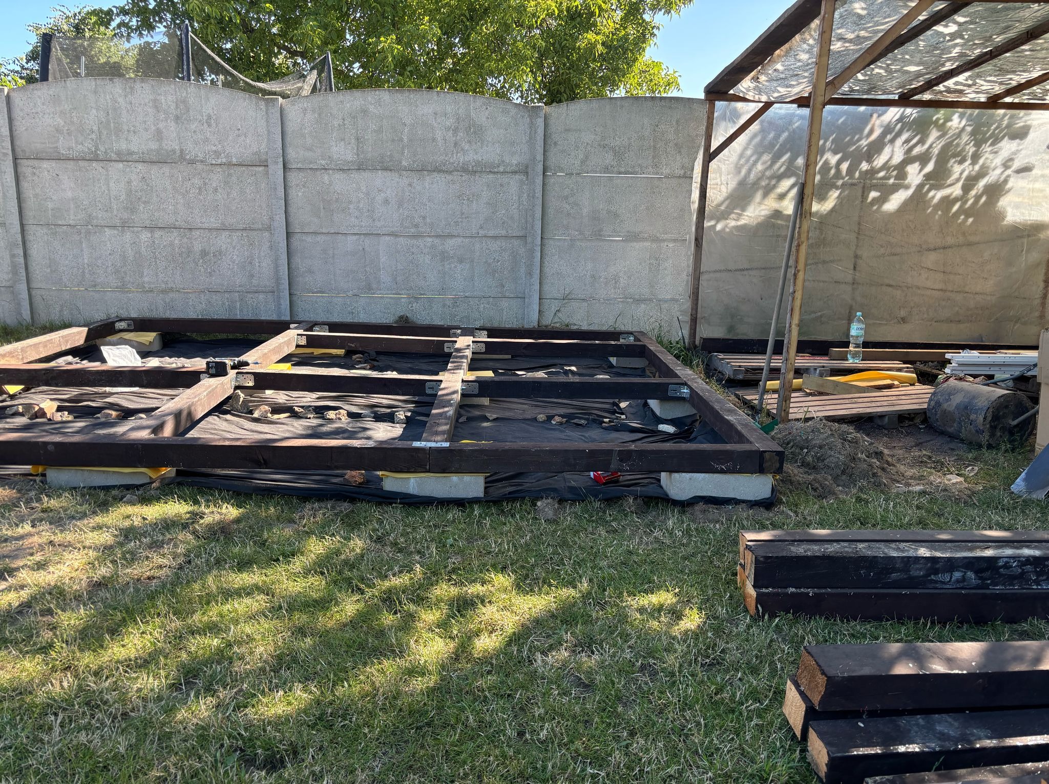 Konstrukcja drewniana tarasu na trawie, z widocznymi elementami mocującymi i czarną folią, na tle betonowego ogrodzenia i zadaszenia. Częściowo zmontowana rama.