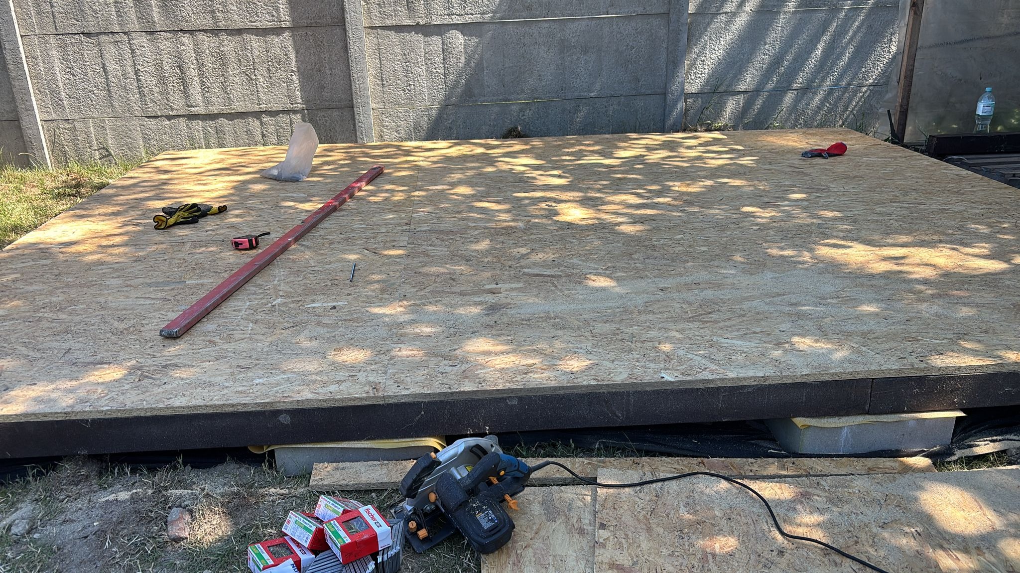 Płyta OSB na legarach, poziomica, rękawice robocze i narzędzia na trawie. Przygotowanie podłoża pod konstrukcję na zewnątrz, widok z góry.