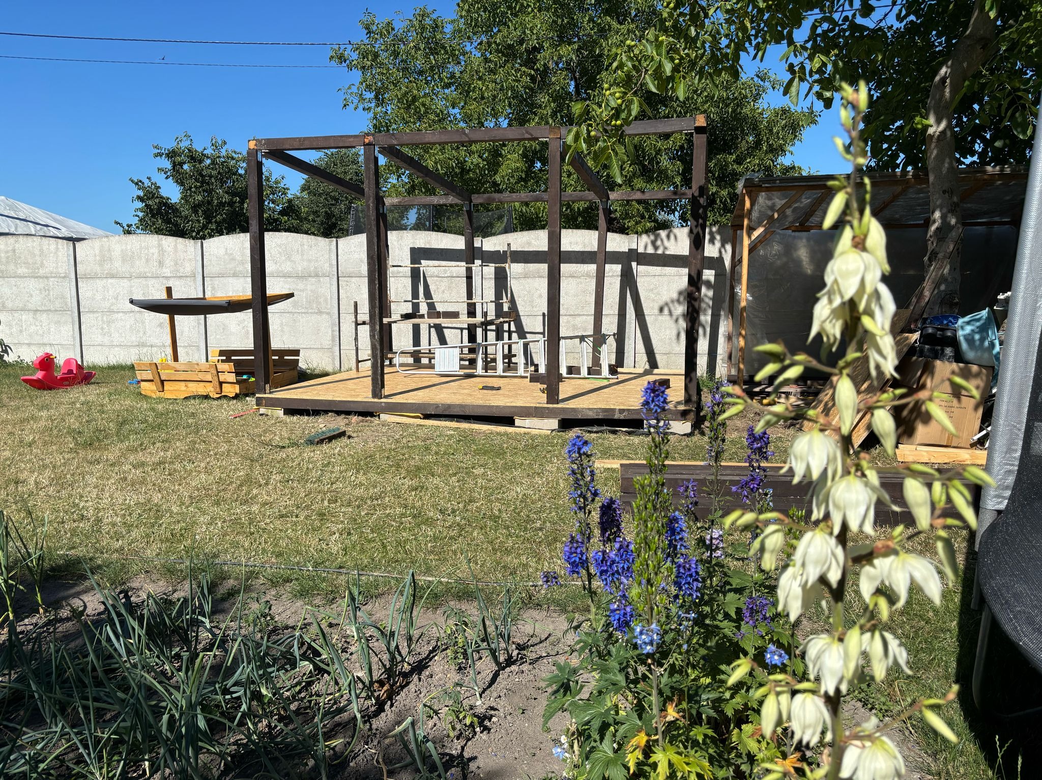 Drewniana konstrukcja altany w ogrodzie, w trakcie montażu. Widoczne narzędzia i drabina. W tle betonowy płot i zieleń. Na pierwszym planie kwiaty.