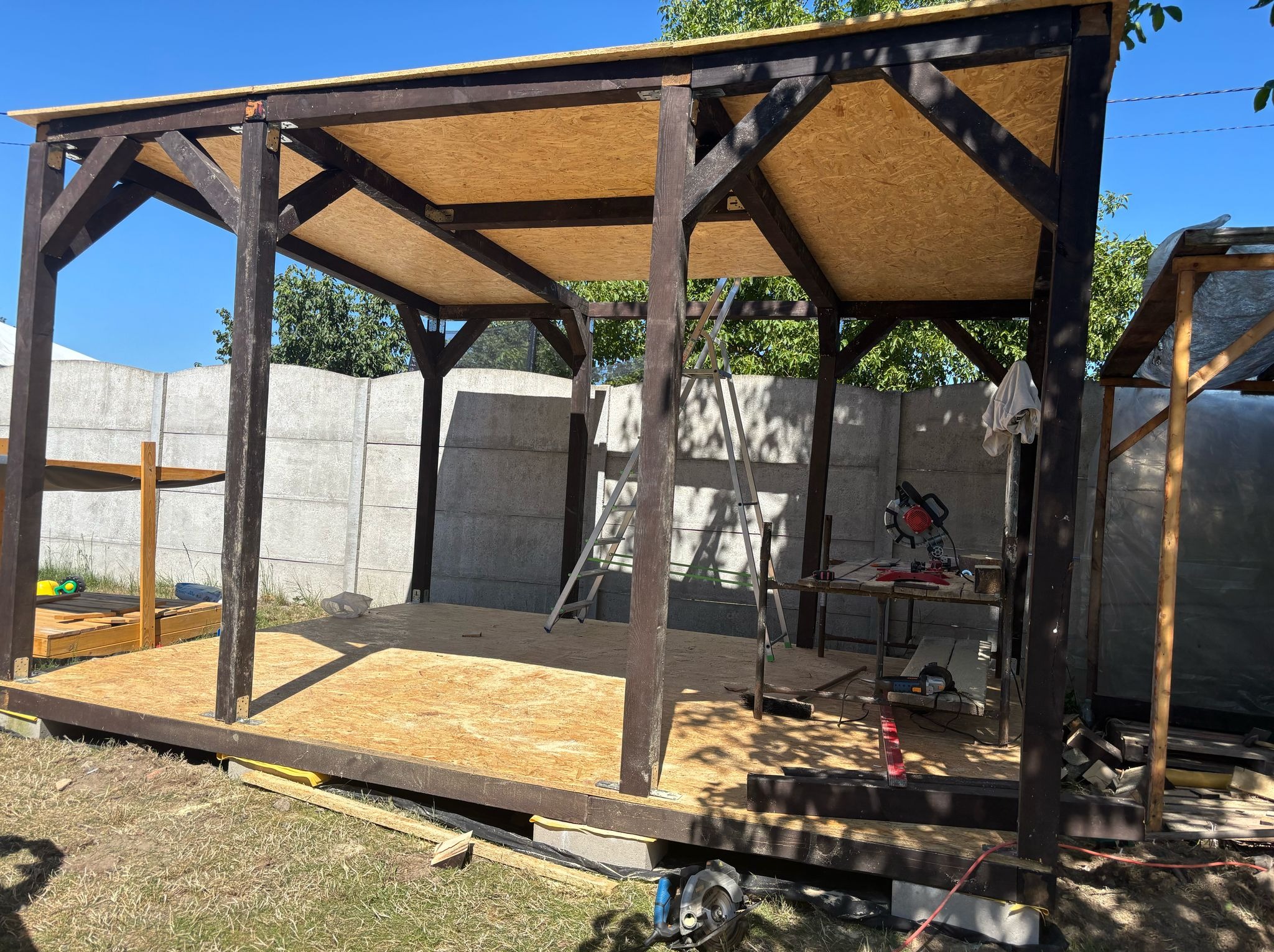 Drewniana konstrukcja altany w trakcie budowy, widoczne narzędzia i drabina, płyty OSB na dachu i podłodze, betonowy płot w tle, słoneczny dzień.