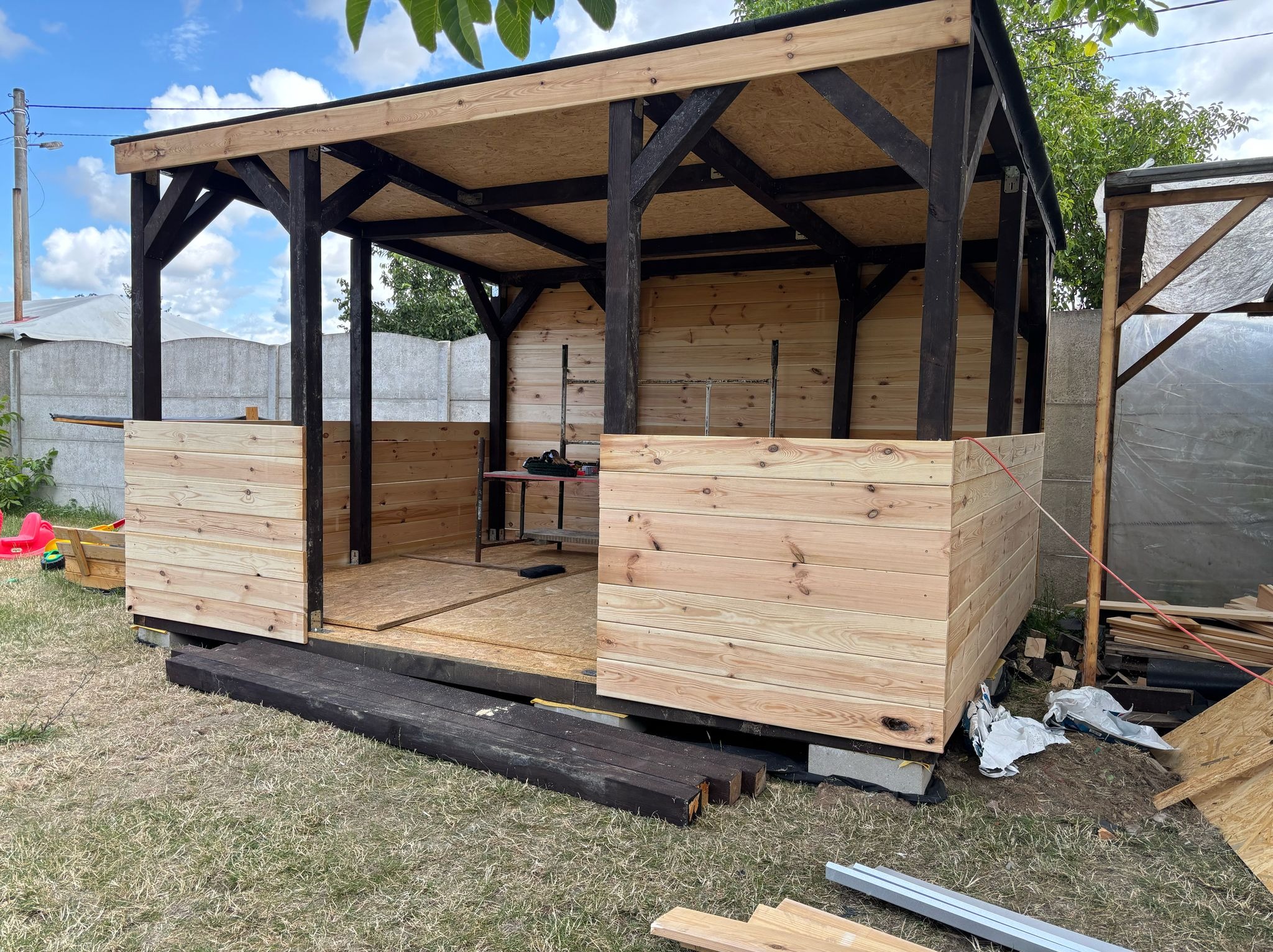 Drewniana altana w trakcie budowy: widoczne deski, konstrukcja z belek, narzędzia w środku. Częściowo ukończone ściany, elementy konstrukcyjne na trawie.