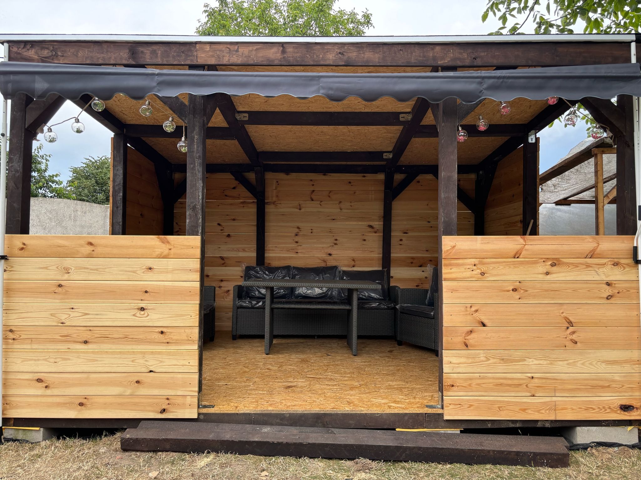 Drewniana altana z meblami ogrodowymi i dekoracyjnymi lampkami. Widoczne deski, konstrukcja dachu i elementy wykończeniowe. Całość w naturalnej kolorystyce drewna.