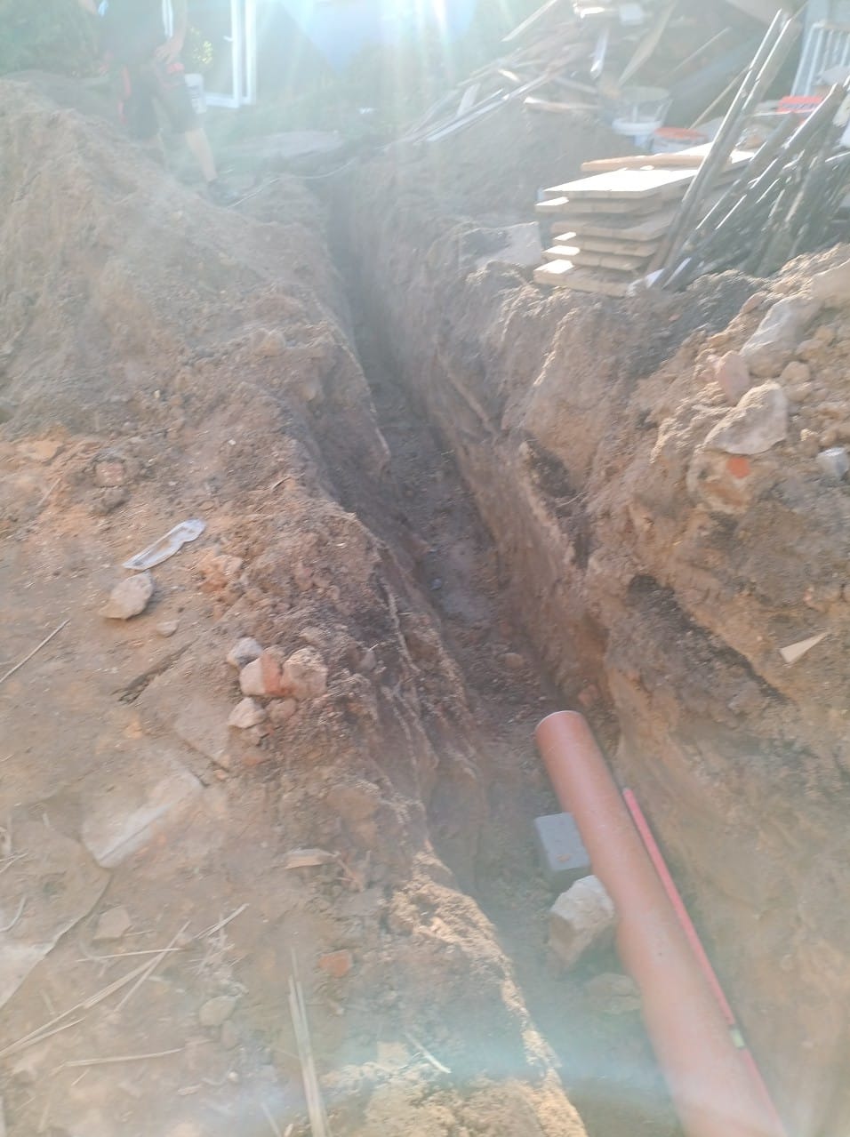 Wykopany wąski rów w ziemi, widoczna częściowo rura z tworzywa sztucznego. W tle deski i inne materiały budowlane. Prace ziemne w słoneczny dzień.