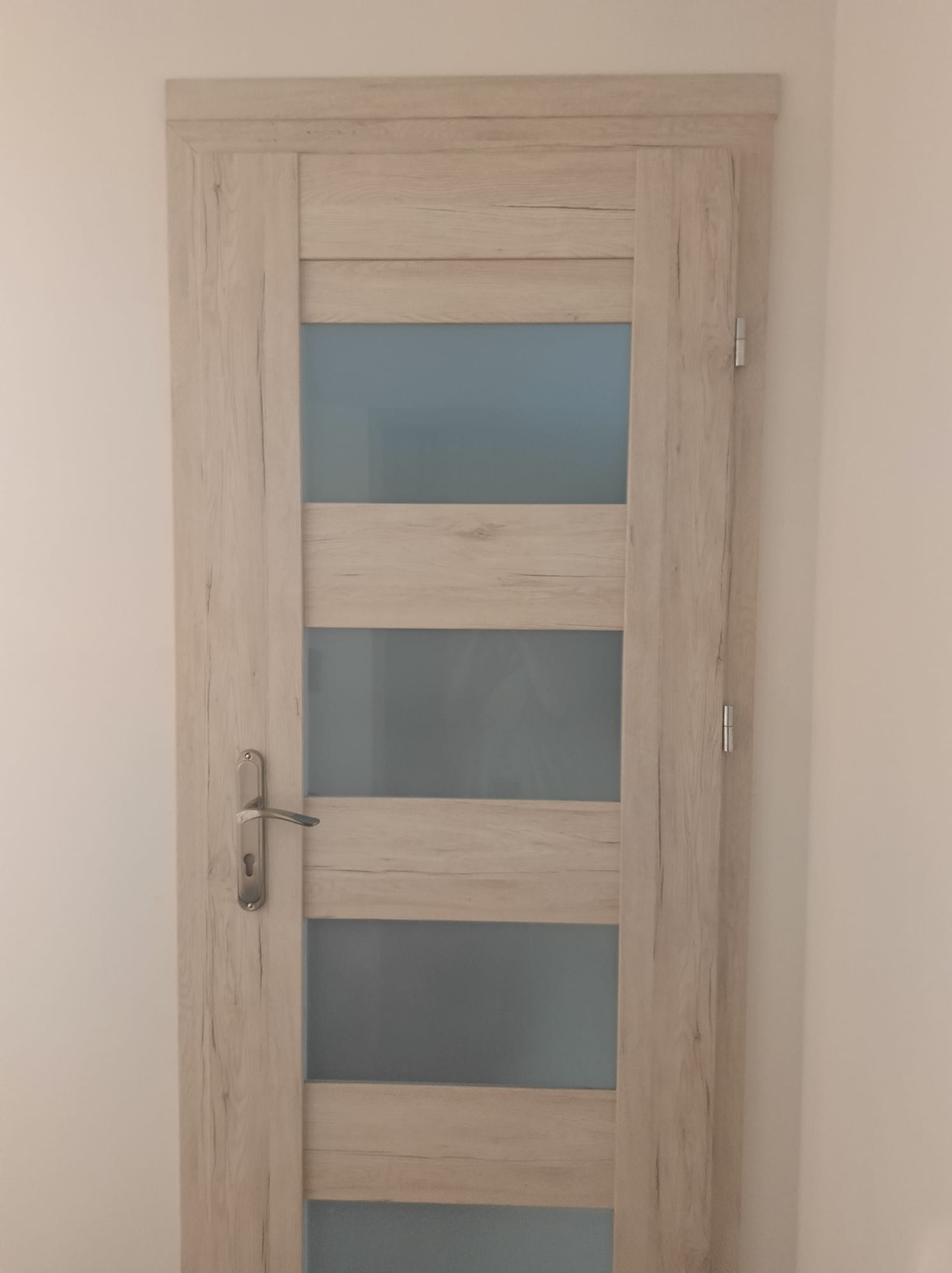 Zamknięte, wewnętrzne drzwi z czterema przeszkleniami i klamką, w odcieniach jasnego drewna, zamontowane w białej ścianie. Widok z przodu, bez osób.
