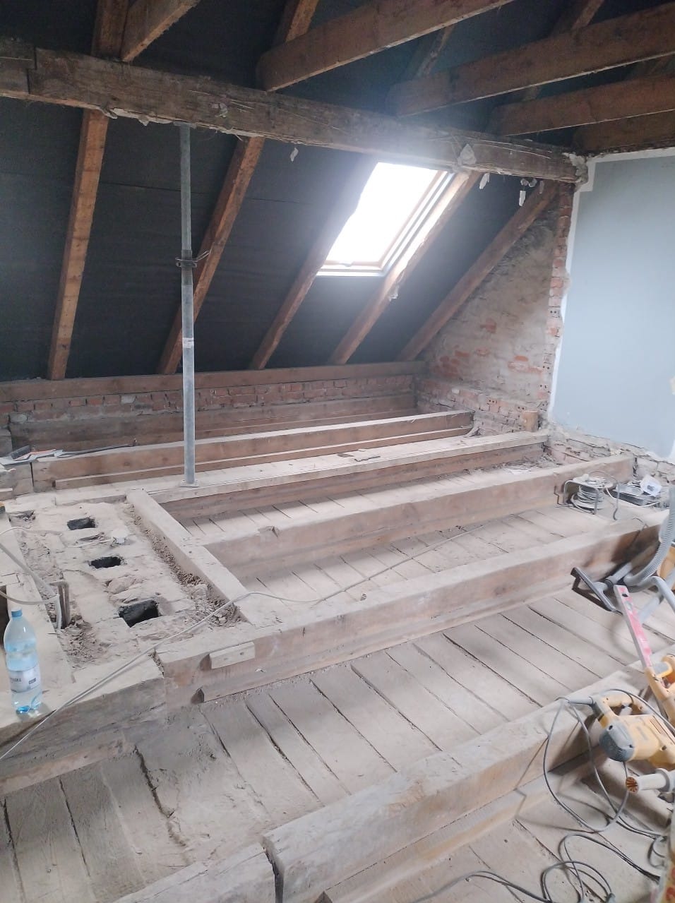 Remont poddasza: drewniana konstrukcja podłogi z widocznymi belkami stropowymi i oknem dachowym. Częściowo odsłonięta cegła i narzędzia budowlane.