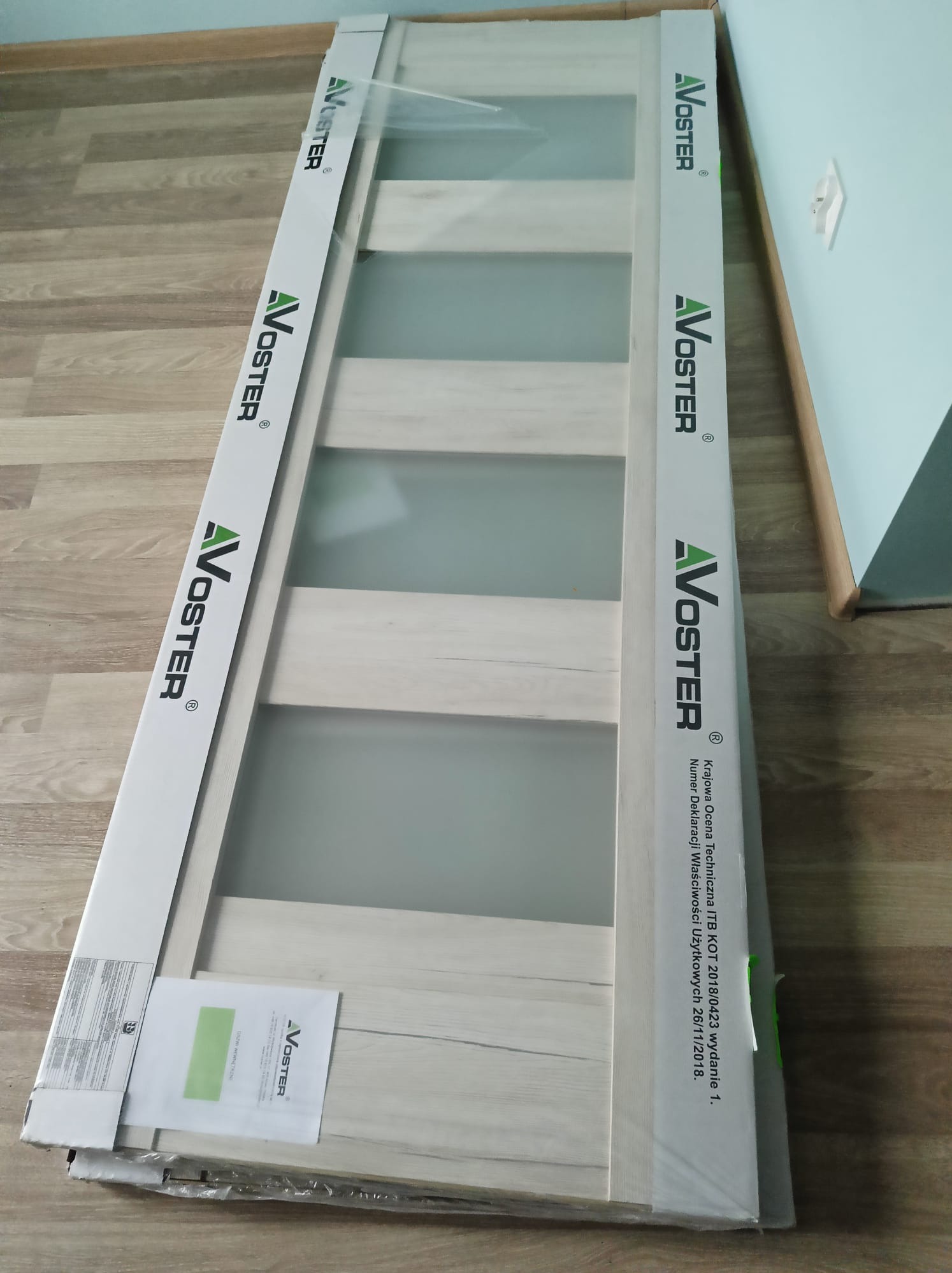 Nowe, zapakowane drzwi wewnętrzne marki Voster z matowym szkłem, leżące na podłodze. Widoczne opakowanie z logo producenta i dokumentacja.