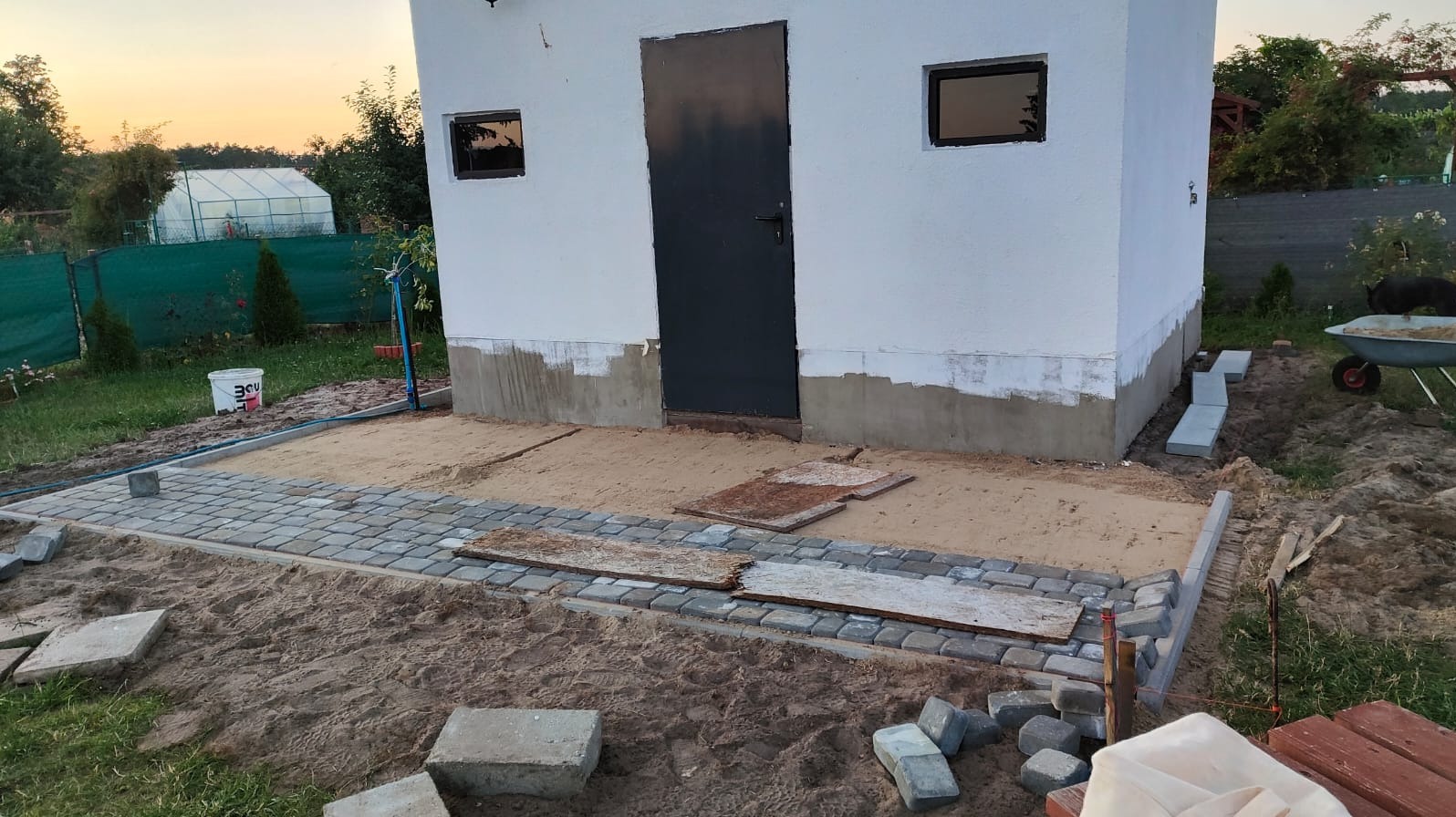 Układanie kostki brukowej przed budynkiem gospodarczym z ciemnymi oknami i drzwiami. Częściowo ułożona kostka, piasek, narzędzia. Widoczny wózek z piaskiem.