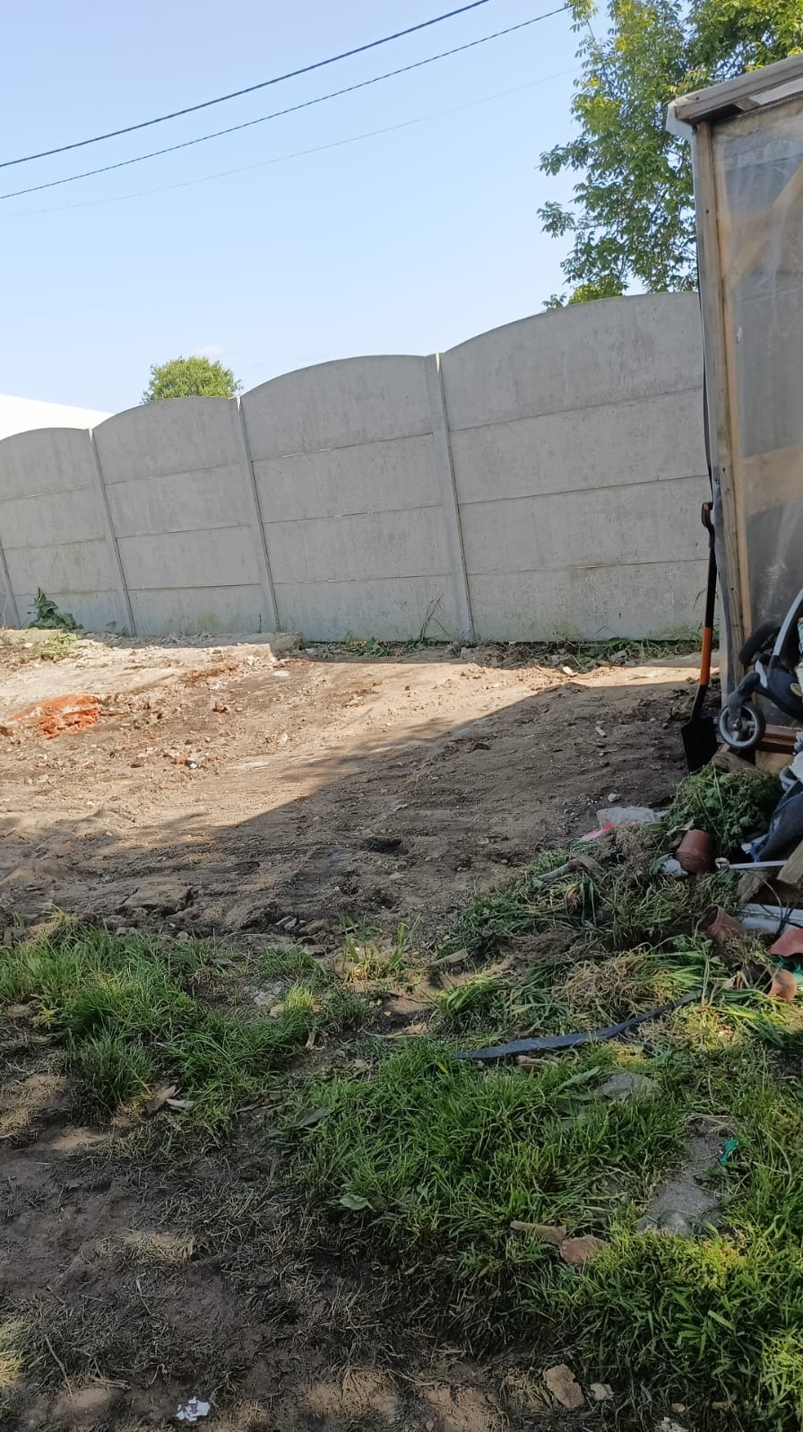 Długi, betonowy płot z zaokrąglonymi górnymi krawędziami, oddzielający teren z ziemią i trawą od nieba. W tle widoczne drzewa i fragment budynku.