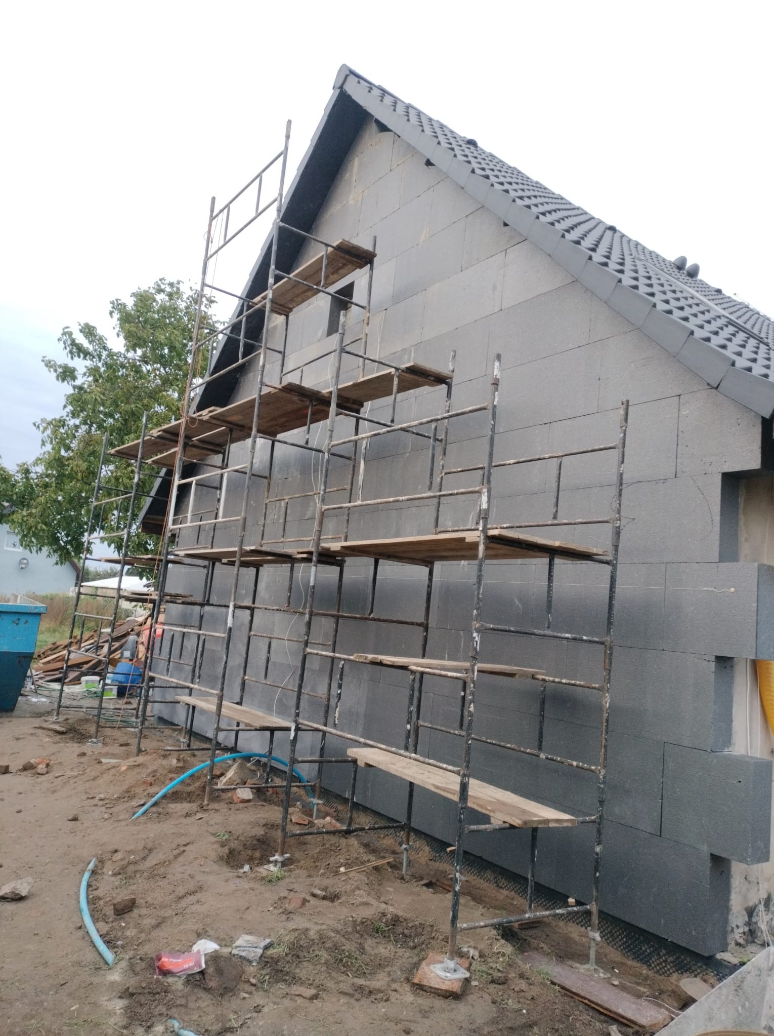 Budynek w trakcie ocieplania styropianem grafitowym, widoczne rusztowanie i fragment dachu z dachówką. Prace remontowe w toku, widok z zewnątrz.