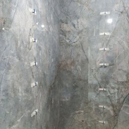 V&MHomePL VOLODYMYR MAIDAN - Ściana w łazience wyłożona szarymi płytkami imitującymi marmur, z widocznymi systemami poziomowania płytek. Widok na narożnik pomieszczenia.