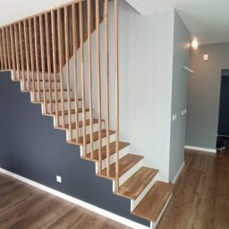 V&MHomePL VOLODYMYR MAIDAN - Nowoczesne schody drewniane z barierką z pionowych listew, łączące dwa poziomy. Ściany pomalowane na szaro i granatowo, podłoga drewniana. Widoczny fragment stołu z akcesoriami malarskimi.