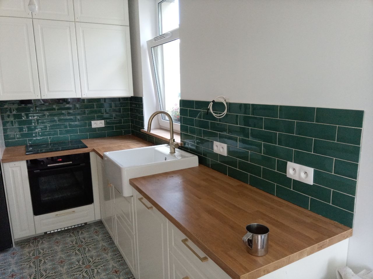 Nowoczesna kuchnia z białymi szafkami, drewnianym blatem i zielonymi płytkami. Uwagę przyciąga złota bateria i wzorzysta podłoga. Widoczny metalowy kubek na blacie.