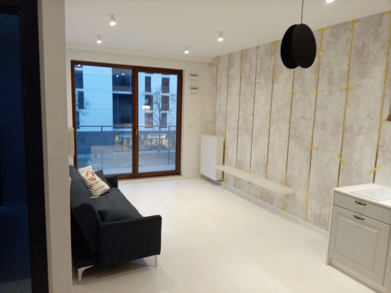 Jasne wnętrze po remoncie: sofa, okno balkonowe, ściana z dekoracyjną tapetą w pionowe pasy. Minimalistyczny design, nowoczesne oświetlenie.