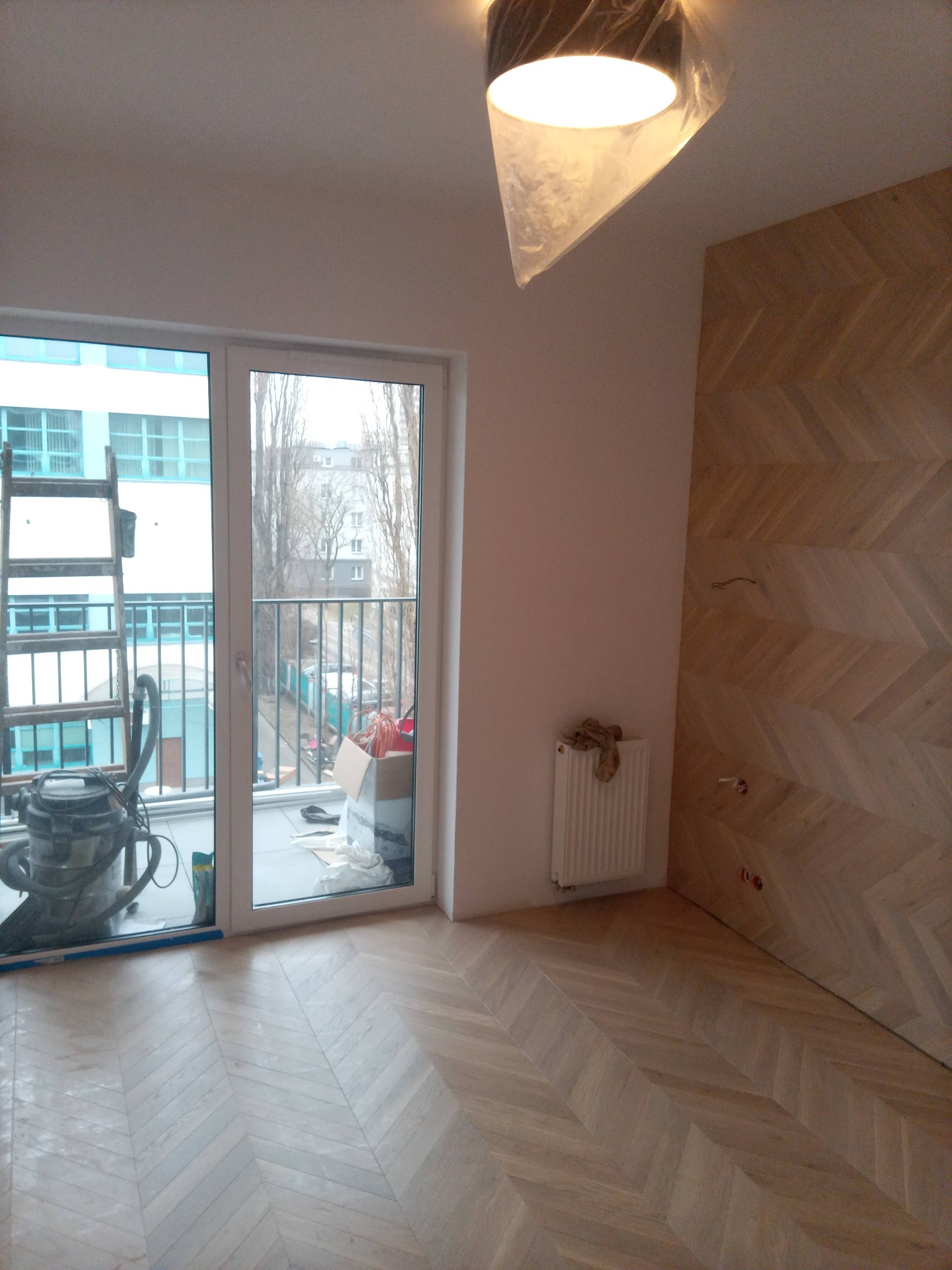 Jasne, nowe parkiety ułożone w jodełkę, ściana z drewnianą okładziną w tym samym wzorze. Widoczny balkon z drabiną i odkurzaczem. Lampa zabezpieczona folią.