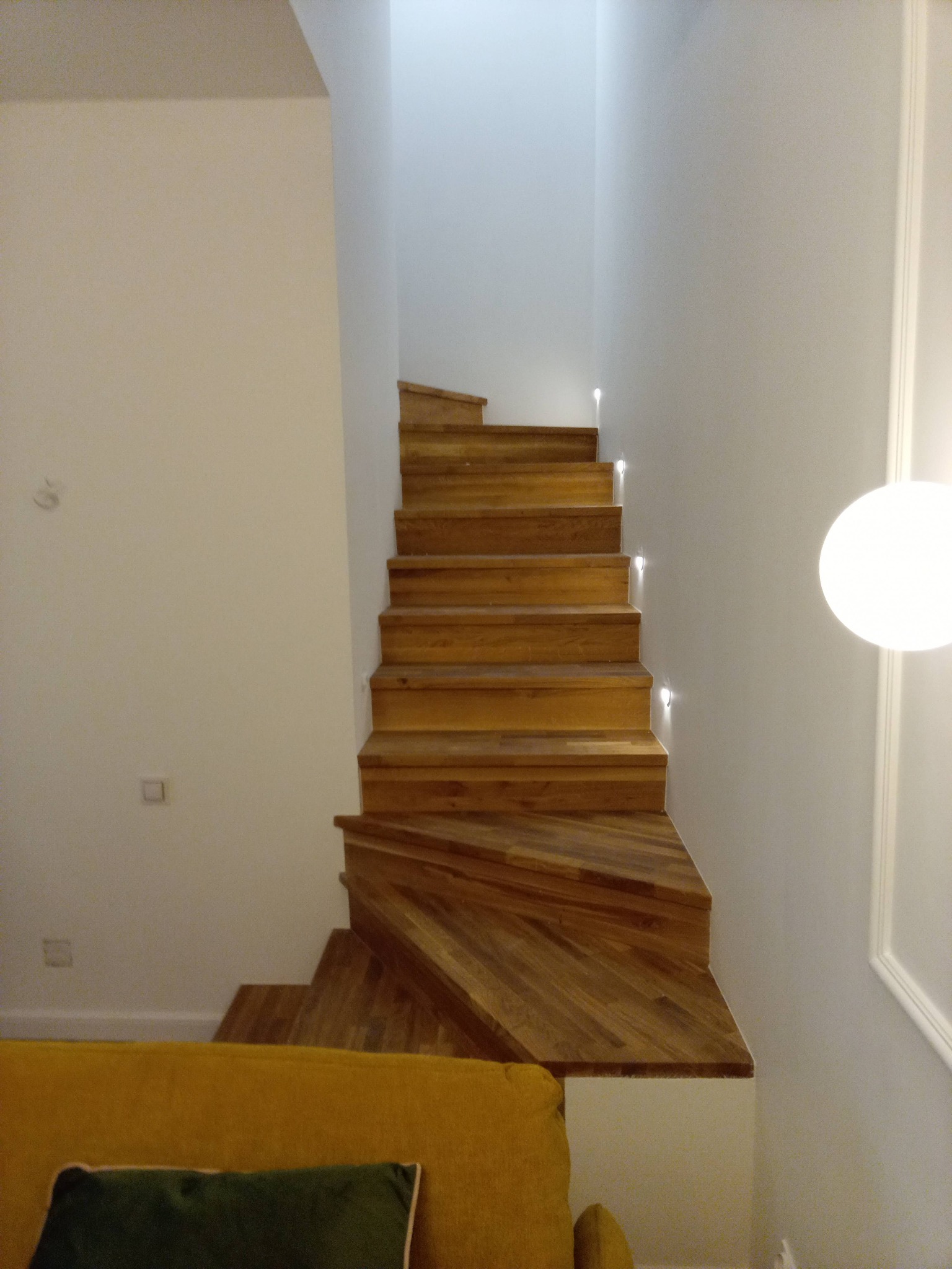 Drewniane schody z oświetleniem punktowym w nowoczesnym wnętrzu, widok z dołu, fragment sofy w kolorze musztardowym w dolnym rogu.