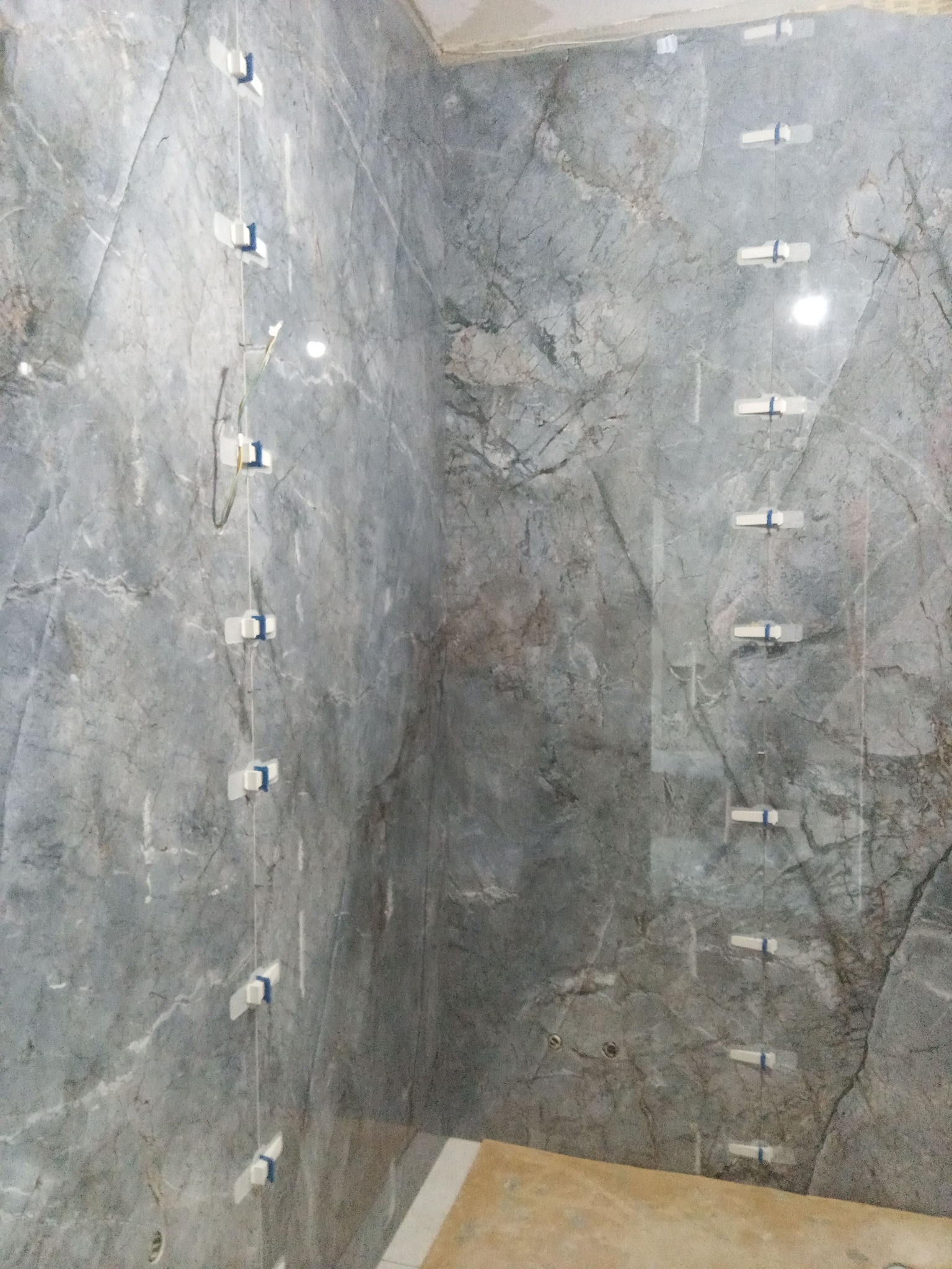 Ściana w łazience wyłożona szarymi płytkami imitującymi marmur, z widocznymi systemami poziomowania płytek. Widok na narożnik pomieszczenia.