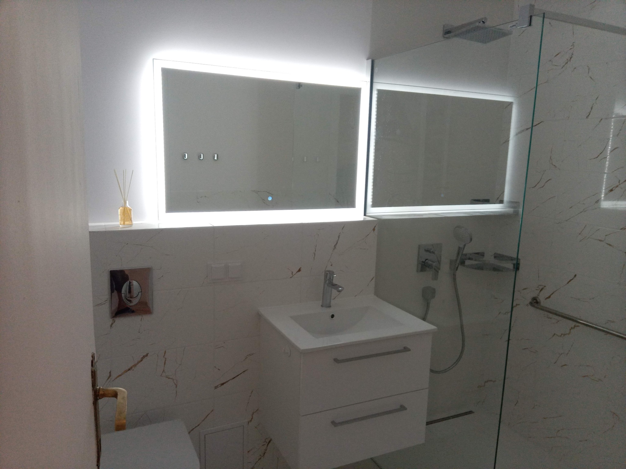 Nowoczesna łazienka z podświetlanym lustrem, białą umywalką z szafką i kabiną prysznicową. Marmurowe płytki na ścianach. Minimalistyczny design.