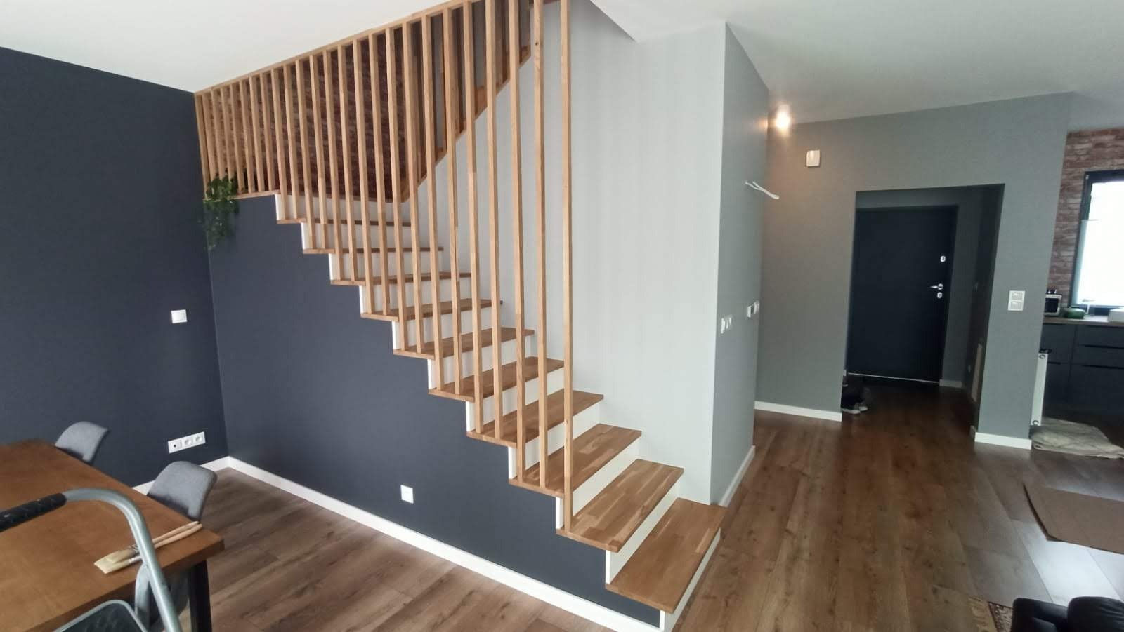 Nowoczesne schody drewniane z barierką z pionowych listew, łączące dwa poziomy. Ściany pomalowane na szaro i granatowo, podłoga drewniana. Widoczny fragment stołu z akcesoriami malarskimi.