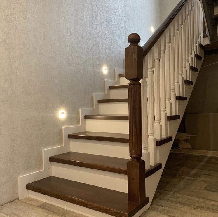 Odnowione schody z drewnianymi stopniami i białymi podstopnicami, oświetlone punktowymi lampami ściennymi. Klasyczny styl z nowoczesnym akcentem.