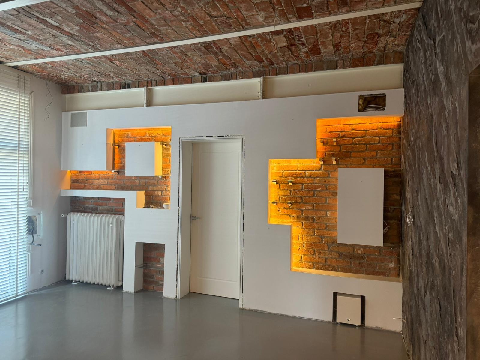 Surowa cegła na suficie i ścianie z wnękami podświetlonymi ciepłym światłem, białe płyty gipsowe, grzejnik i drzwi. Wnętrze w trakcie wykańczania.