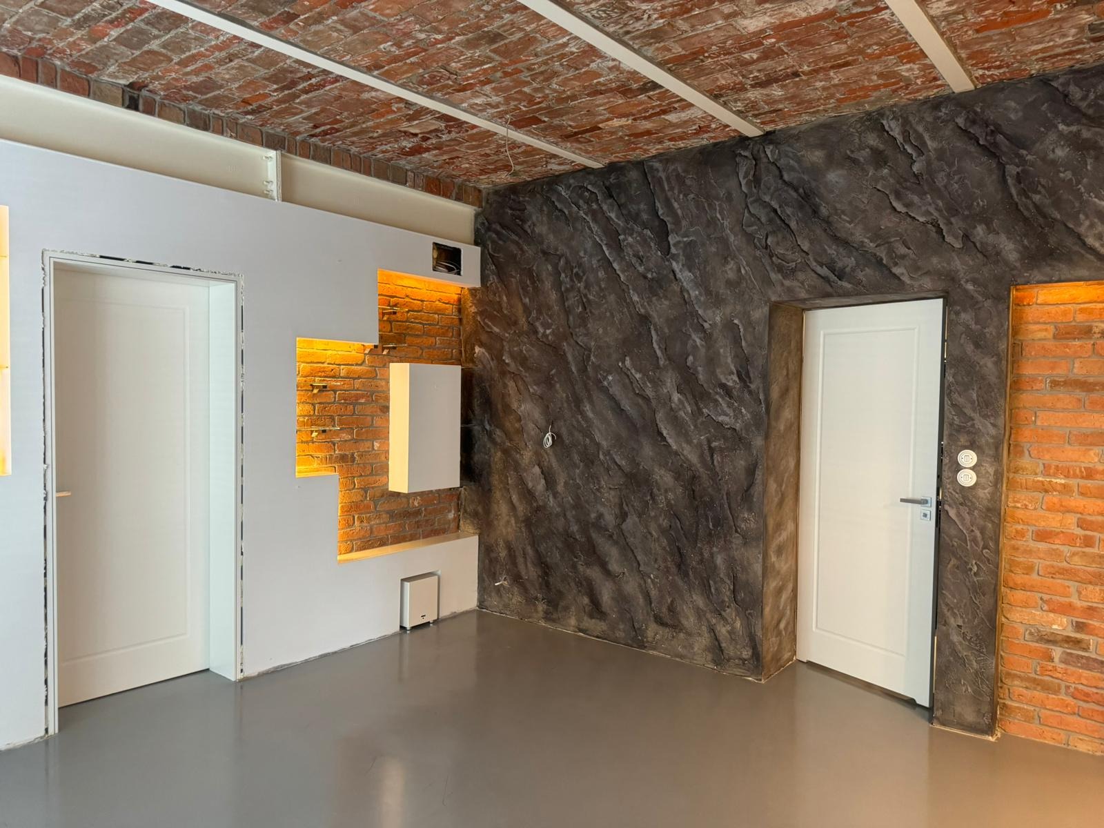 Loftowe wnętrze po remoncie: ceglany sufit, ściana z efektem kamienia, białe drzwi i oświetlenie LED wnęk. Nowoczesny design z surowymi elementami.