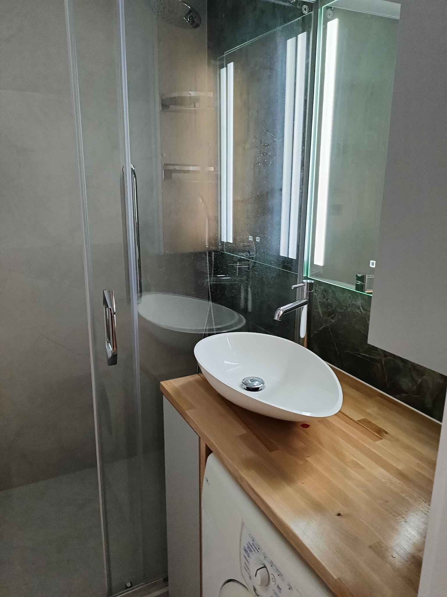 Nowoczesna łazienka po remoncie: kabina prysznicowa ze szklanymi drzwiami, umywalka nablatowa na drewnianym blacie, podświetlane lustro. Minimalistyczny design.