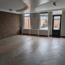Henryk Kosiewski - Jasny parkiet ułożony w jodełkę francuską w przestronnym pokoju z ceglaną ścianą, białymi grzejnikami pod oknami i designerskim sufitem podwieszanym.