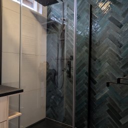 Henryk Kosiewski - Nowoczesna kabina prysznicowa z czarnym brodzikiem i armaturą, ściana wyłożona błyszczącymi, zielono-niebieskimi płytkami w jodełkę. Widoczny fragment szafki z drewnianymi półkami.
