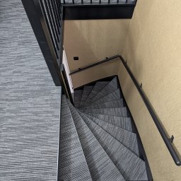 Henryk Kosiewski - Spiralne schody z szarą wykładziną dywanową, widok z góry. Czarne metalowe balustrady i beżowa ściana. Nietypowy kąt ujęcia podkreśla geometrię schodów.