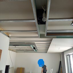 Henryk Kosiewski - Montaż sufitu podwieszanego z płyt kartonowo-gipsowych. Widoczne metalowe profile, częściowo zamontowane płyty i instalacja oświetleniowa. W tle drabina i materiały budowlane.