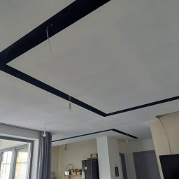 Henryk Kosiewski - Biały sufit z czarnymi, geometrycznymi pasami dekoracyjnymi, widoczne żarówki na kablach, fragment okna i wyposażenia wnętrza. Minimalistyczny design.