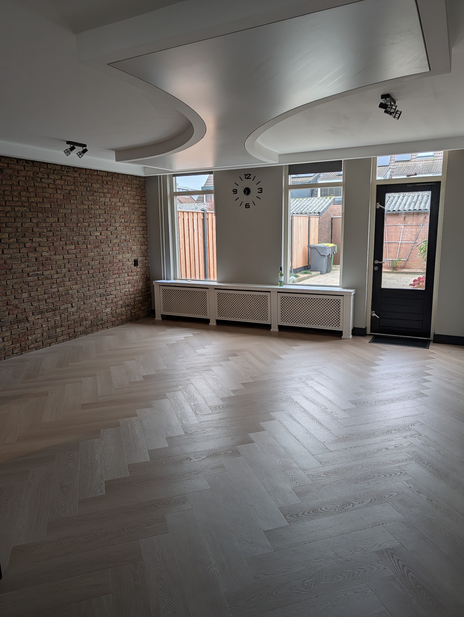 Jasny parkiet ułożony w jodełkę francuską w przestronnym pokoju z ceglaną ścianą, białymi grzejnikami pod oknami i designerskim sufitem podwieszanym.