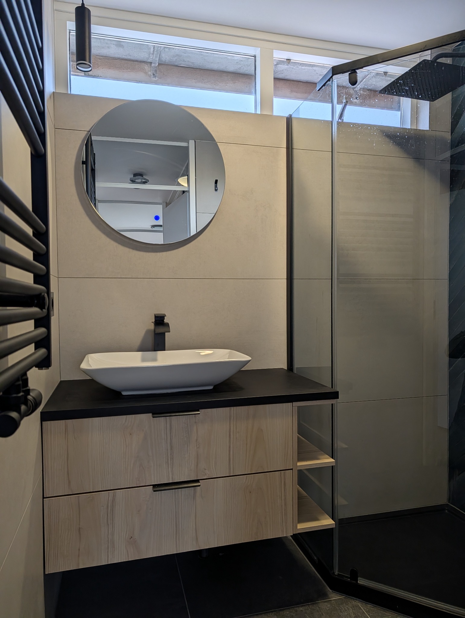 Nowoczesna łazienka z umywalką nablatową, okrągłym lustrem i kabiną prysznicową. Uwagę zwraca drewniana szafka pod umywalką z czarnym blatem.